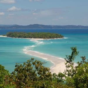 Ein erhöhter Blick auf eine weiße Sandbank, die zu einer üppigen, baumbestandenen Insel führt, umgeben von türkisem und tiefblauem Wasser.