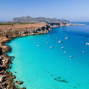 Una vista aerea di diverse imbarcazioni bianche e catamarani ancorati in una baia turchese, accanto a una costa rocciosa, sotto un cielo azzurro e limpido.