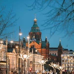 Una via della città decorata con luci festive porta verso una grande cattedrale in mattoni rossi con cupole verdi al crepuscolo.