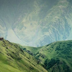 Un monastero in pietra con un campanile si erge in cima a una verde collina erbosa, sullo sfondo di imponenti montagne nebbiose.