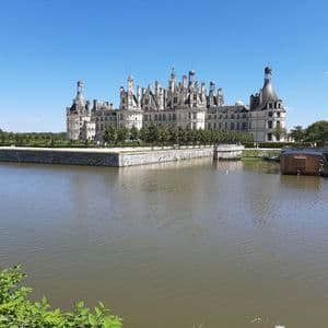 Un grand château de pierre blanche, orné de nombreuses tourelles, se dresse derrière de larges douves, sous un ciel bleu dégagé.