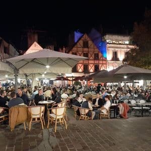 Un café en plein air animé sur une place pavée la nuit, avec des gens dînant à des tables sous de grands parasols.