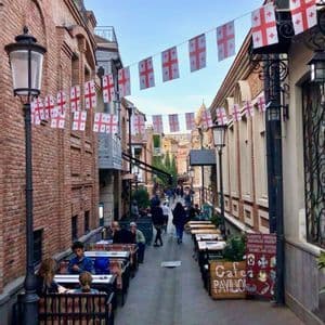 Una strada stretta con edifici in mattoni è fiancheggiata da tavolini di caffè all'aperto e decorata con festoni di bandiere.