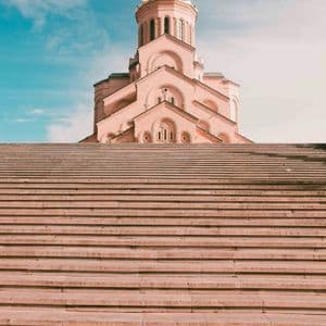 Un'ampia scalinata in pietra conduce a una maestosa cattedrale dalle tonalità rosa, sullo sfondo di un cielo azzurro vibrante.