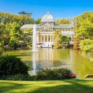 Un ornato palazzo di vetro si erge di fronte a un lago con una grande fontana, circondato da rigogliosi alberi verdi in un parco sotto un cielo azzurro e limpido.