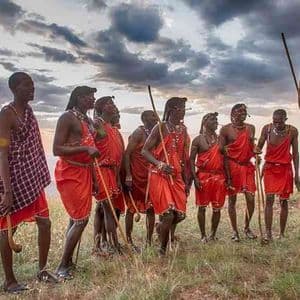 Un grupo de hombres masái con vestimenta roja tradicional en una colina de hierba, con uno realizando un salto alto al atardecer.