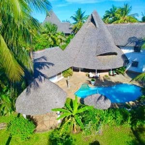 Una vista aérea de una villa tropical con techos de paja y una piscina, rodeada de exuberantes palmeras verdes.