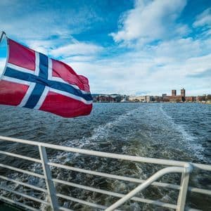 Un drapeau norvégien flotte à l'arrière d'un bateau, laissant un sillage dans l'eau avec un horizon urbain en arrière-plan.