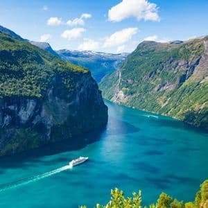 Vue aérienne d'un bateau de croisière blanc naviguant dans un fjord turquoise, entouré de montagnes vertes escarpées sous un ciel bleu.