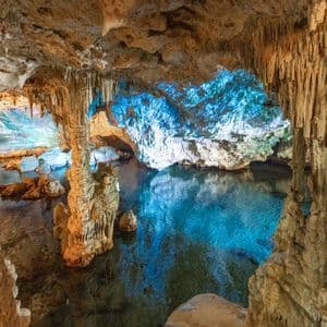 Stalattiti e stalagmiti all'interno di una vasta caverna con una limpida piscina sotterranea illuminata da luci blu e gialle.