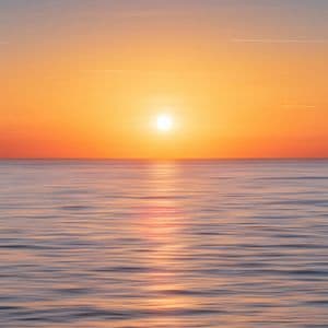 Il sole è basso all'orizzonte, diffondendo un bagliore arancione e giallo su un mare calmo con dolci increspature.