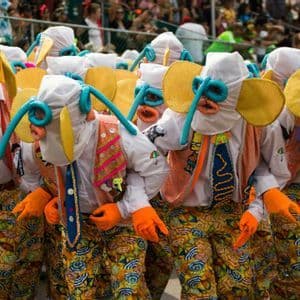 Un gruppo di persone in costumi vivaci con maschere a forma di elefante e pantaloni a motivi geometrici si esibisce durante una parata di strada.