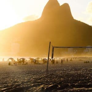 Una rete da pallavolo si erge su una spiaggia affollata al tramonto, con una grande montagna che si staglia contro la luce dorata.