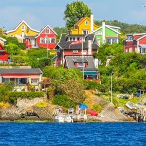 Des maisons en bois colorées sont construites sur une colline rocheuse verdoyante, au bord d'une étendue d'eau d'un bleu profond, avec des bateaux amarrés en contrebas.