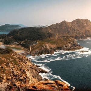 Una vista en picada de una península rocosa y arbolada que incluye un istmo de arena y una pequeña laguna, rodeada por el intenso azul del mar.
