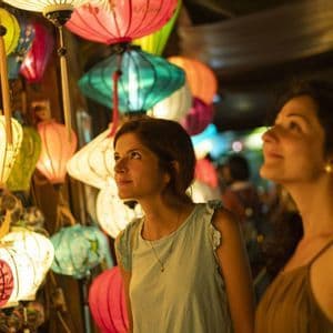 Deux femmes se tiennent à un étal de marché la nuit, regardant un étalage de lanternes en papier colorées et illuminées.