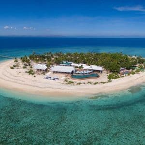 Vista aerea di una piccola isola tropicale con un resort, circondata da una spiaggia di sabbia bianca e acqua turchese cristallina.