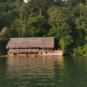 Un lodge en bois à deux étages, avec un toit de chaume, se dresse sur l'eau au pied d'une colline dense et boisée.