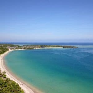 Veduta aerea di una spiaggia curva di sabbia bianca, che costeggia acqua turchese e una lussureggiante foresta verde, sotto un cielo sereno.