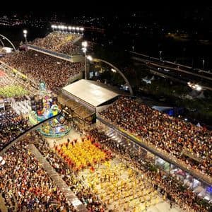 Vista aérea de un vibrante desfile de carnaval nocturno con artistas disfrazados y carrozas elaboradas, rodeado de grandes multitudes en las gradas.
