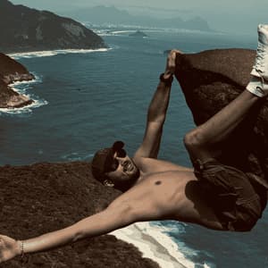 Un homme souriant suspendu à un rocher pour une photo, avec une vue plongeante sur une côte avec des plages et des collines luxuriantes.