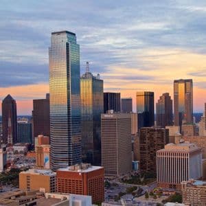 A dense city skyline with numerous skyscrapers reflecting the warm light of a sunset under a partly cloudy sky.