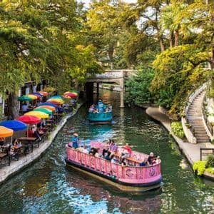 Two colorful tour boats with a WeRoad group trip cruise down a canal lined with lush trees and cafes with rainbow umbrellas.