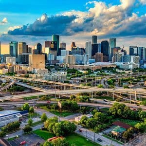 An aerial view of a modern city skyline with multiple highway interchanges in the foreground under a cloudy blue sky.