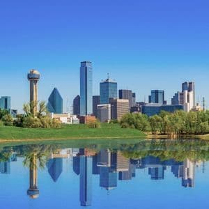 A modern city skyline with skyscrapers reflected perfectly in calm water, with a green grassy bank in the foreground.