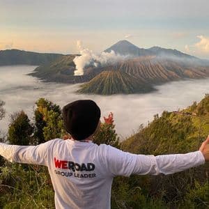 Un chef de groupe WeRoad, vu de dos, se tient les bras tendus, surplombant une vallée de nuages et des cratères volcaniques fumants à l'aube.