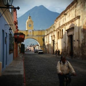 Une rue pavée bordée de bâtiments coloniaux mène à une arche jaune surmontée d'une tour d'horloge, avec un grand volcan en arrière-plan.