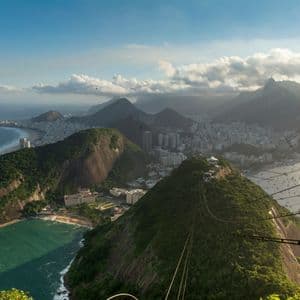 Un teleférico se mueve a lo largo de sus cables sobre una exuberante montaña verde, con vistas a una extensa ciudad costera y una bahía con barcos.