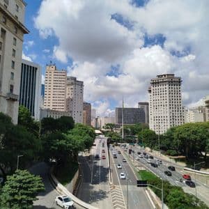 Vista elevada de una autopista de varios carriles que atraviesa una ciudad con rascacielos y exuberantes árboles bajo un cielo azul nublado.