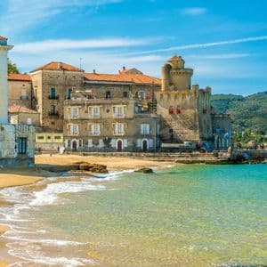 Un pueblo costero con edificios históricos y un castillo se asienta en una playa de arena junto a aguas cristalinas de color turquesa bajo un cielo azul.