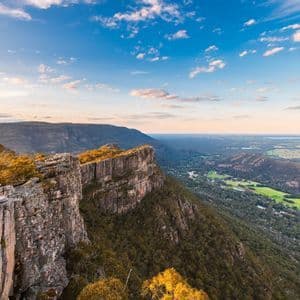 Ein Panoramablick von einer felsigen Klippe, der ein bewaldetes Tal und weite Ebenen unter einem blauen Himmel mit Wolken bei Sonnenuntergang zeigt.