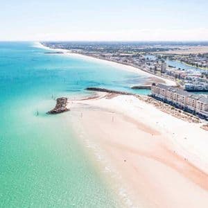 Luftaufnahme eines langen weißen Sandstrandes und türkisfarbenen Ozeans neben einer Küstenstadt mit einem Yachthafen.