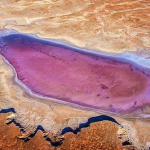 Eine Luftaufnahme eines länglichen rosa Sees, umgeben von einer trockenen, kargen Landschaft in Braun- und Orangetönen.