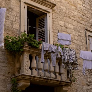 Wäsche hängt an einer Wäscheleine vor einem Steinbalkon mit Topfpflanzen und einem geöffneten Fenster an einer rustikalen Gebäudefassade.