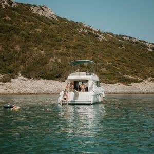 Eine WeRoad-Gruppenreise entspannt auf einer weißen Yacht, die in einem ruhigen Meer ankert, mit einigen Leuten, die neben einer grünen, felsigen Küste schwimmen.
