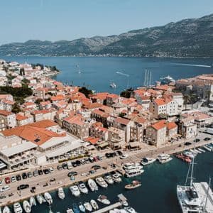 Eine Luftaufnahme einer Küstenstadt mit orangefarbenen Ziegeldächern, einem Hafen voller Boote und Bergen über dem blauen Wasser.