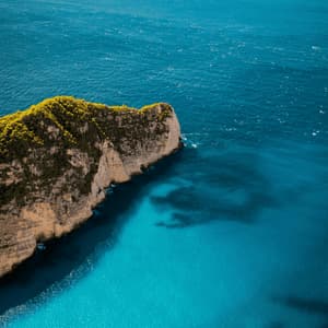 Luftaufnahme einer felsigen, grün bewachsenen Klippe, die in ein türkisfarbenes und tiefblaues Meer ragt.