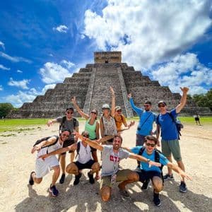 A WeRoad group trip of friends posing for a photo in front of a large stone step pyramid under a bright blue sky.
