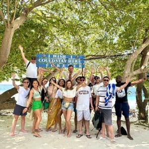 Un groupe WeRoad pose ensemble pour une photo sur une plage de sable sous un grand arbre avec un panneau bleu.