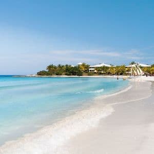 Ein weißer Sandstrand mit türkisfarbenem Wasser, das sanft ans Ufer plätschert. Im Hintergrund sind Palmen, Segelboote und Resortgebäude zu sehen.