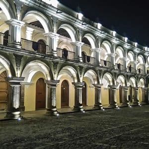 Un largo edificio colonial de dos plantas con una arcada de arcos iluminada con luces ascendentes en una plaza adoquinada por la noche.