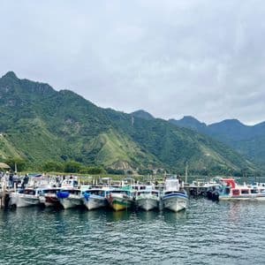 Una flota de pequeños barcos está atracada en un muelle de madera en un lago, con grandes montañas verdes elevándose al fondo bajo un cielo nublado.