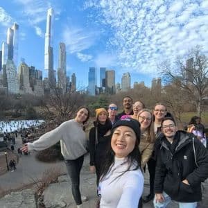 A woman takes a selfie of her WeRoad group trip posing on a large rock, with a city skyline and an ice rink in the background.