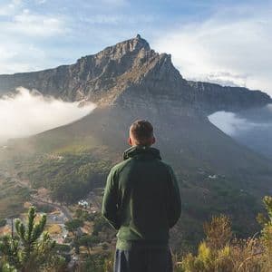 Eine Person in einem grünen Kapuzenpullover steht auf einem Aussichtspunkt und blickt auf einen großen, felsigen Berggipfel, der teilweise von Wolken bedeckt ist.