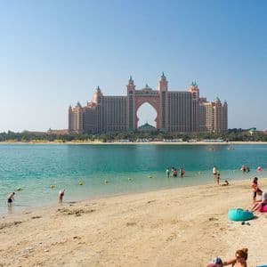 Le persone si rilassano e nuotano su una spiaggia assolata, con un grande hotel ornato che presenta un arco prominente, affacciato sull'acqua cristallina.