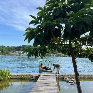 Un hombre ata una pequeña lancha motora a un muelle de madera en un lago, con un árbol verde exuberante en primer plano y colinas al fondo.